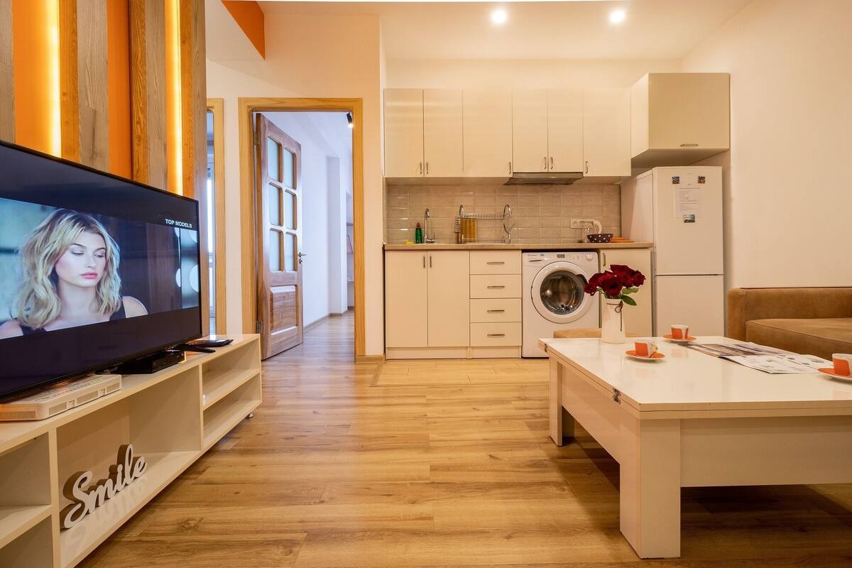 A modern kitchen with a washing machine, a dining area with a table and chairs, and a TV on a stand.
