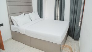 Una cama con cabecera gris, ropa de cama blanca y cortinas grises.
