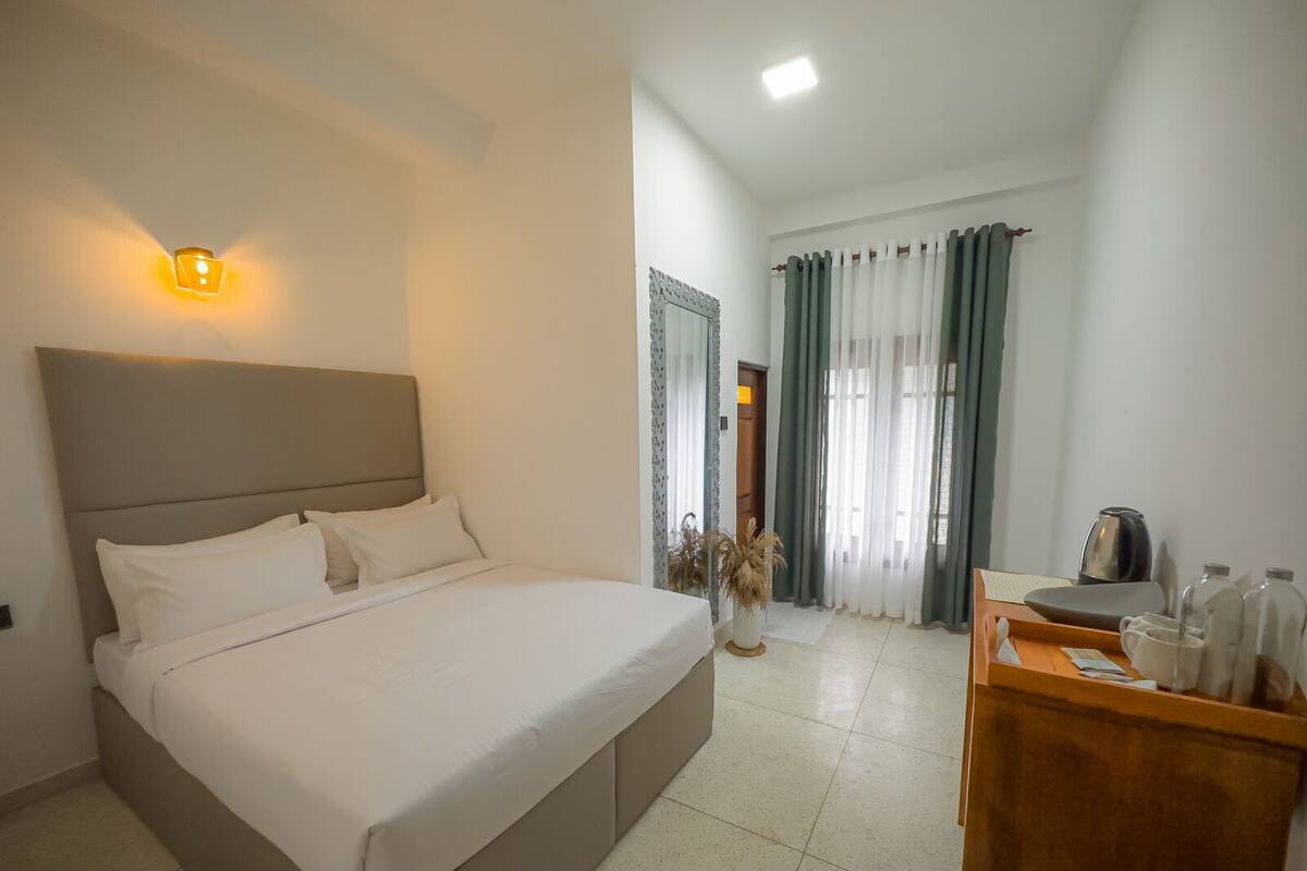 A hotel room with a large bed, a bedside table with a kettle, and a view of the hallway with curtains.