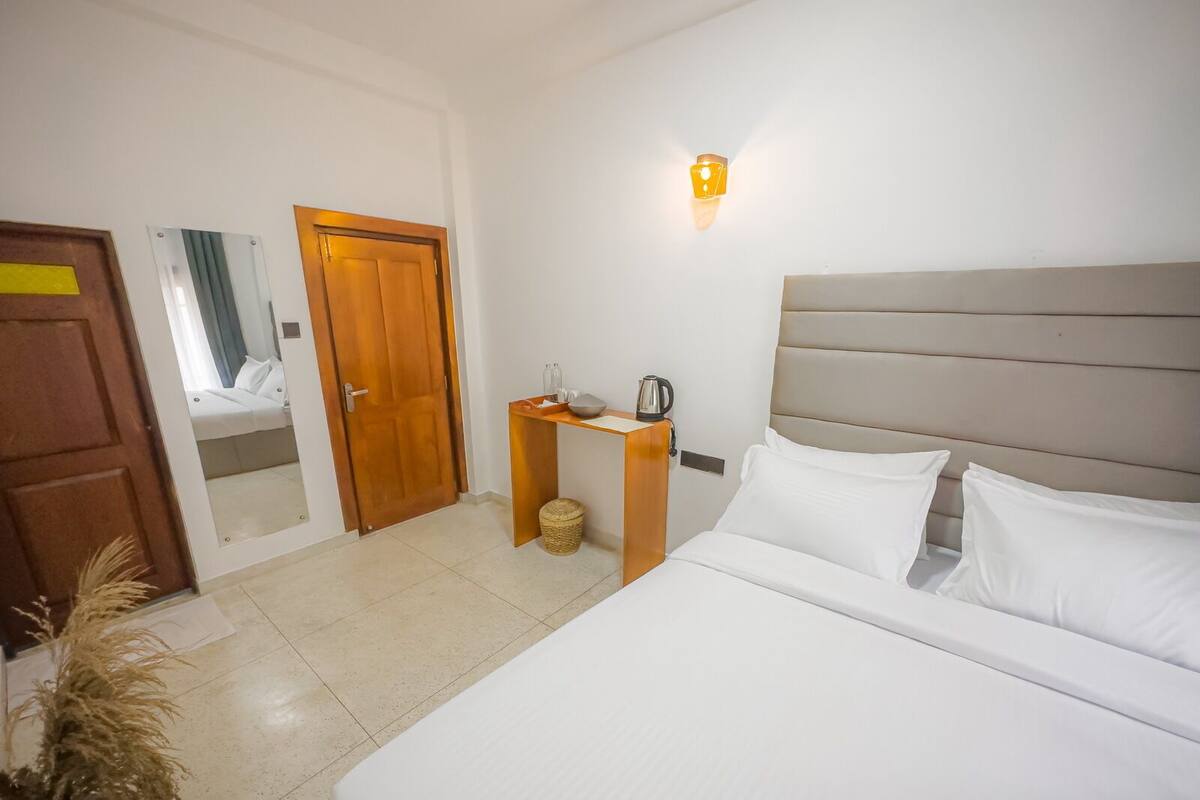 A hotel room with a bed, a nightstand, a wall-mounted light, and a door leading to another room.