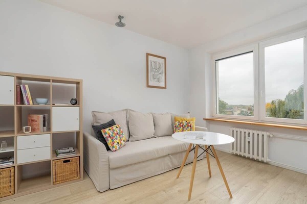 A bright living space with a gray sofa, wooden shelves, a small coffee table, and large windows allowing natural light.