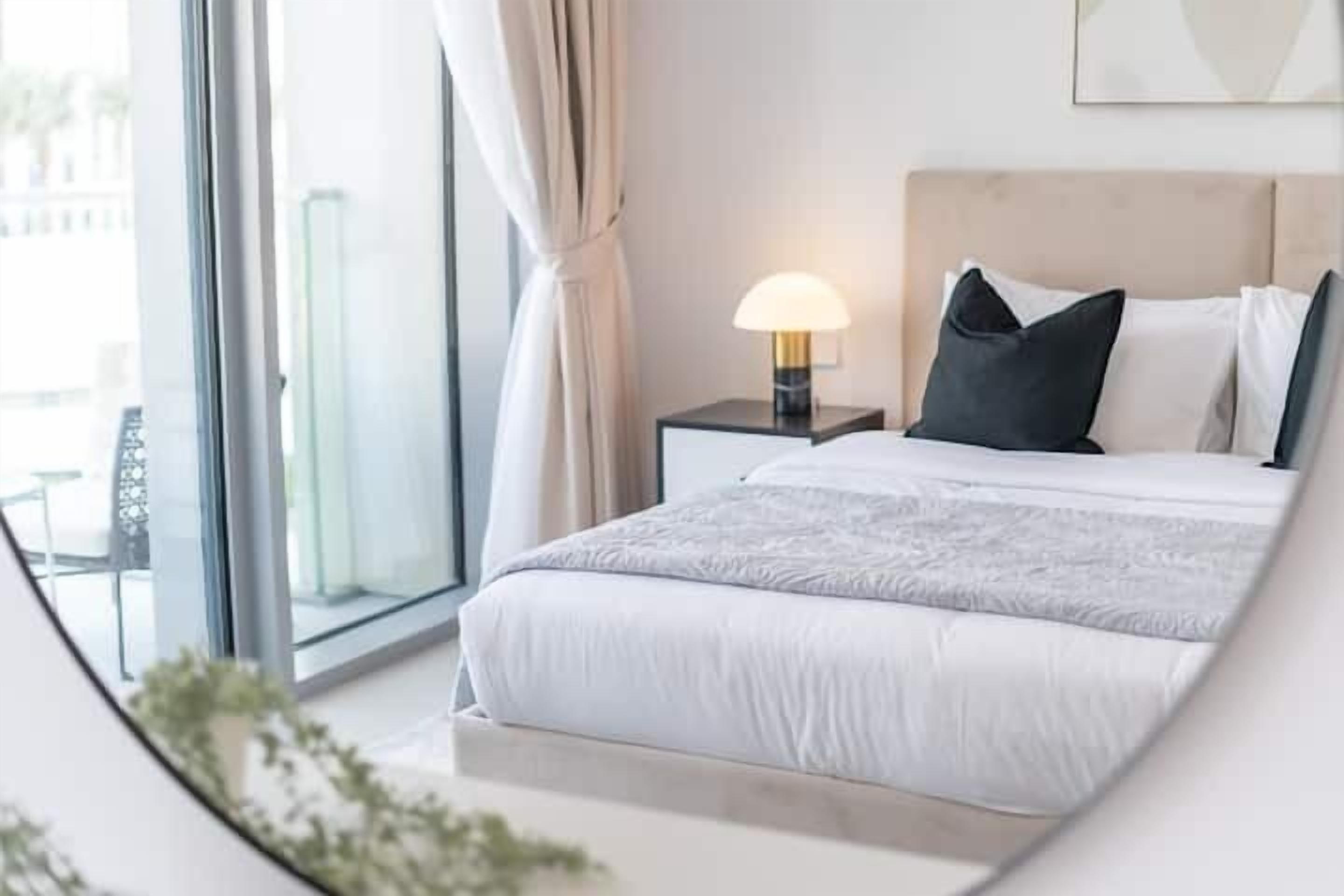 A modern hotel room with a large bed, a bedside table with a lamp, and a view through a large window.
