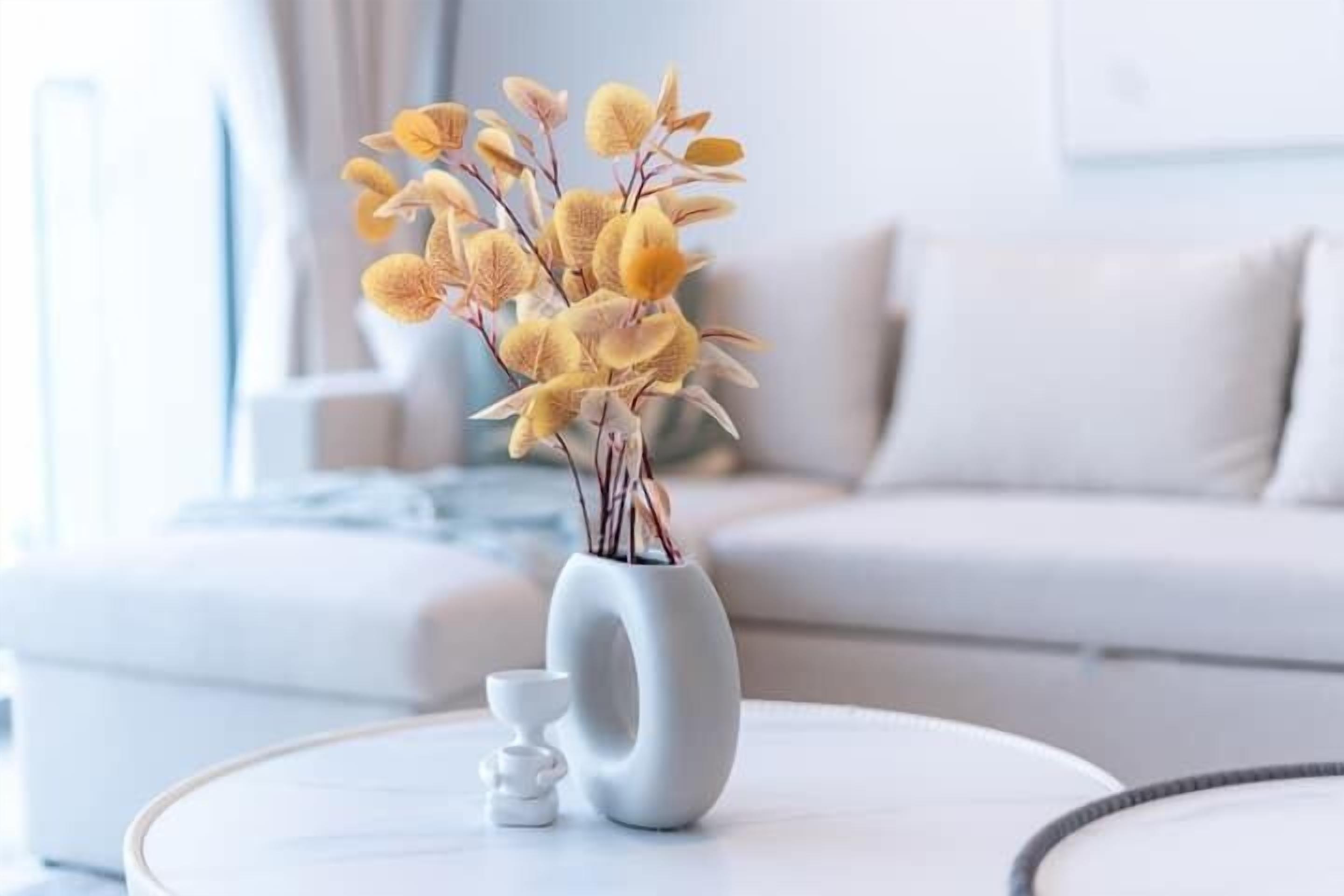 Eine weiße Vase mit gelben Blumen auf einem weißen runden Tisch in einem modernen Wohnzimmer mit einem weißen Sofa und einem weißen Couchtisch.