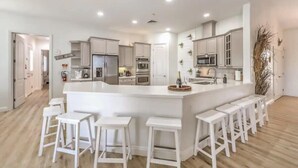 A modern kitchen with white cabinets, a central island with bar stools, and a visible refrigerator. - Lux 5BR Villa w Pool, Spa, RV Parking, Sleeps 12 (Henderson)