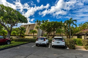 A parking lot with two cars, trees, and a building with a modern design. - Maui Kamaole #A-104 Front Row, Ocean View, Across Kamaole Bch III, Full A/C (Kihei)
