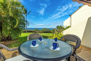 A round table with four blue cups, surrounded by wicker chairs, set on a patio with a view of the ocean and palm trees. - Maui Kamaole #A-104 Front Row, Ocean View, Across Kamaole Bch III, Full A/C (Kihei)