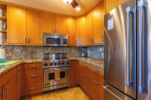 A kitchen with wooden cabinets, a stainless steel stove, a microwave, and a refrigerator. - Maui Kamaole #A-104 Front Row, Ocean View, Across Kamaole Bch III, Full A/C (Kihei)