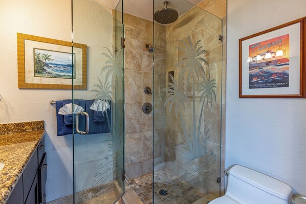 A bathroom with a glass-enclosed shower, a toilet, a framed picture of a beach, and a framed Hawaii poster. - Maui Kamaole #A-104 Front Row, Ocean View, Across Kamaole Bch III, Full A/C (Kihei)
