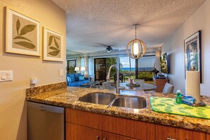 A kitchen with granite countertops, a double sink, and wooden cabinets. There is a ceiling fan, a hanging light fixture, and artwork on the walls. - Maui Kamaole #A-104 Front Row, Ocean View, Across Kamaole Bch III, Full A/C (Kihei)