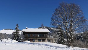 Een houten chalet met een met sneeuw bedekte dak, omringd door besneeuwde bomen en bergen op de achtergrond.