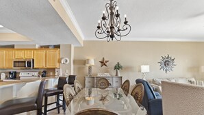 Un vaste espace de vie comprenant un grand salon avec une table à manger en verre entourée de chaises, un coin cuisine doté de meubles en bois et un lustre.