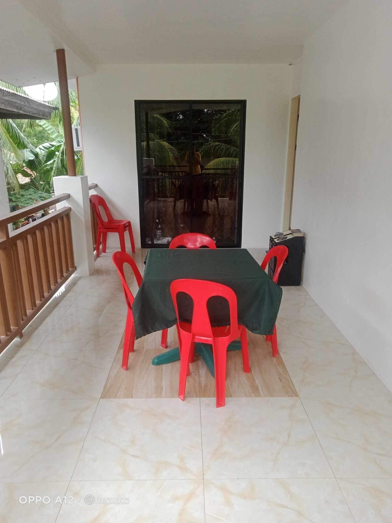 Un comedor con una mesa dispuesta para cuatro personas, rodeado de sillas rojas, un mantel verde y vistas a un jardín a través de un ventanal.