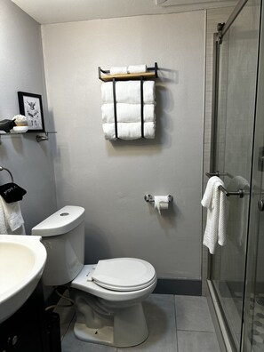 A bathroom with a toilet, sink, and shower. There is a towel rack with towels and a framed picture on the wall.