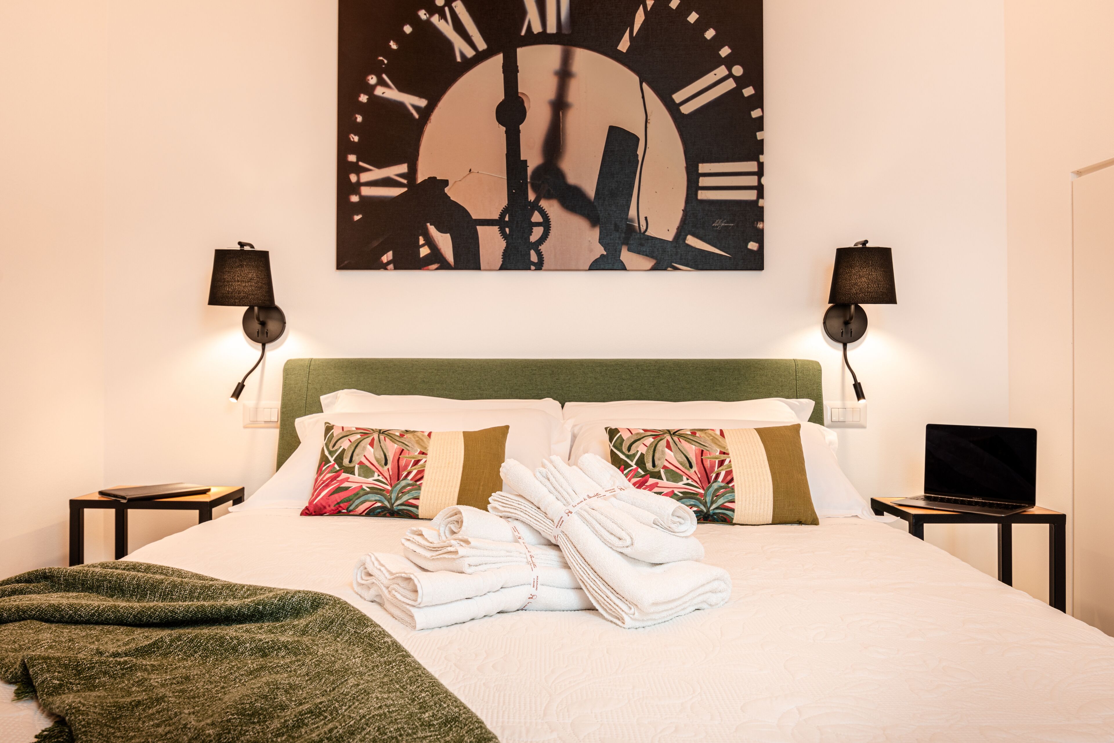 Un lit bien fait, avec des serviettes pliées, un plaid vert et deux oreillers aux motifs tropicaux. Une grande horloge murale avec des chiffres romains est accrochée au-dessus du lit.