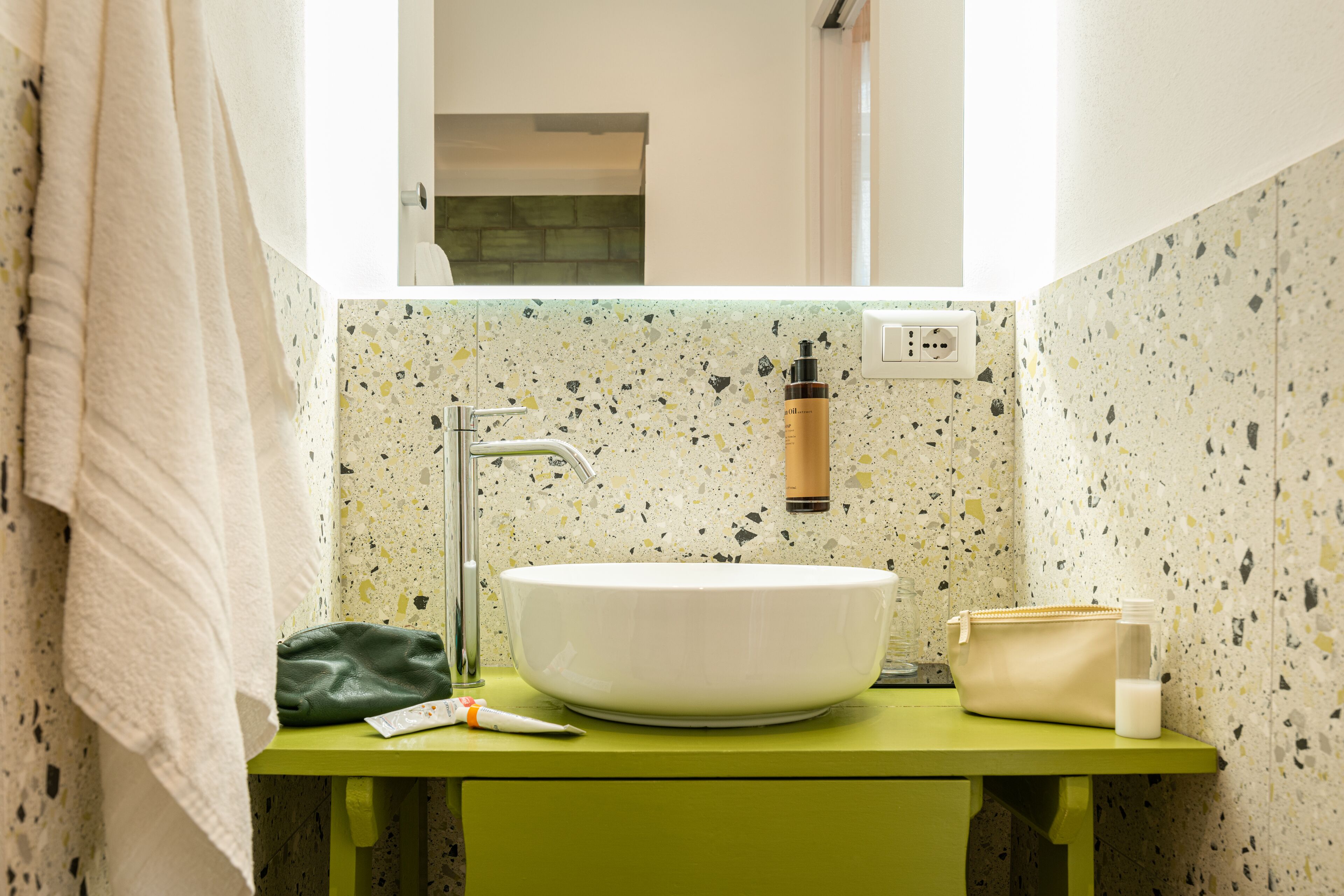 Un baño con un lavabo verde, una pileta blanca, una toalla, una bolsa verde y artículos de aseo.