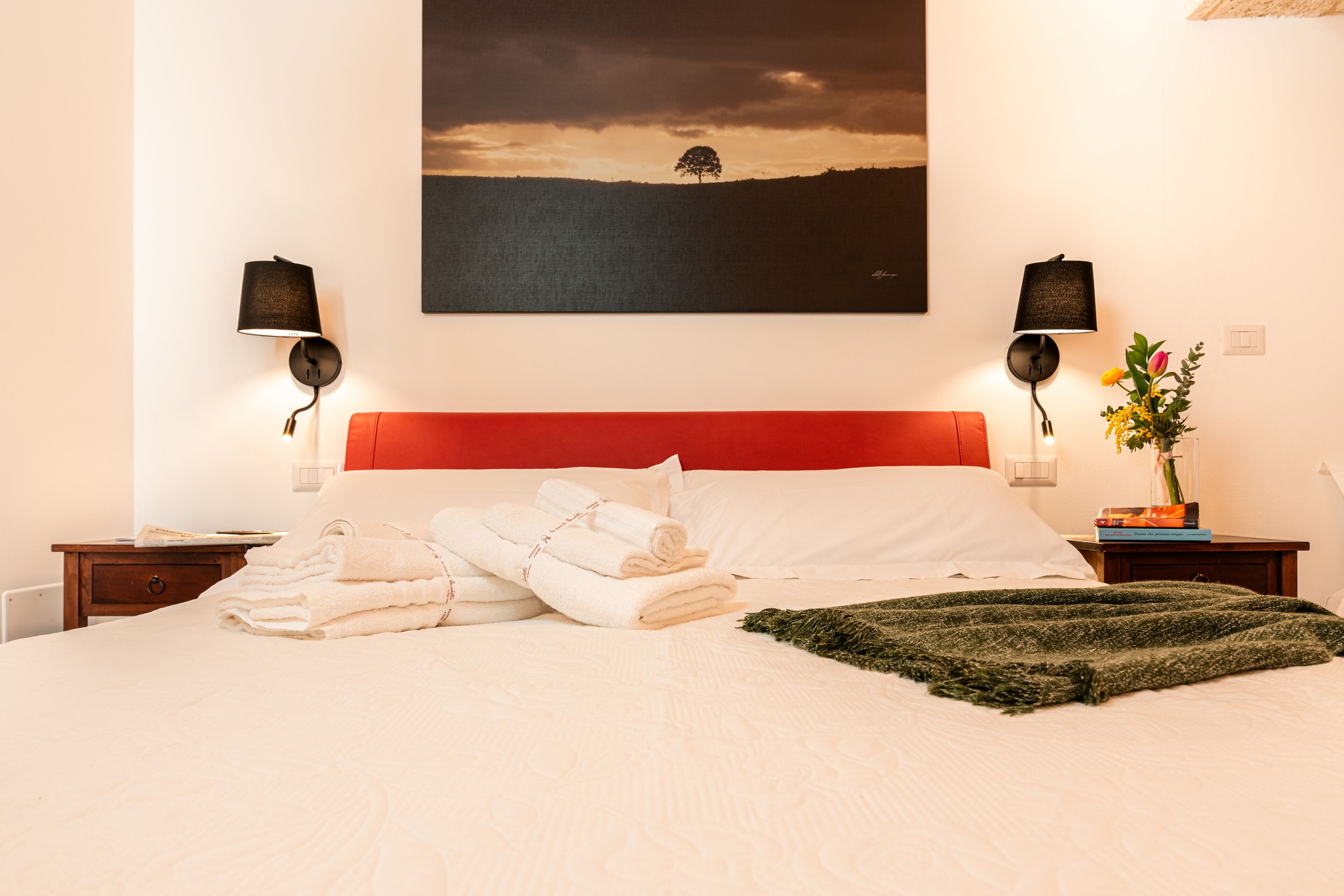 A neatly made bed with white linens and a green throw, a red headboard, and a painting of a tree on the wall.