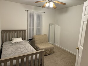 Ein Schlafzimmer mit Bett, Sessel, Deckenventilator, Fenster mit durchsichtigen Vorhängen und einer Tür.