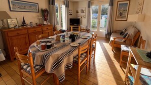 Zona pranzo con tavolo in legno, sedie e tovaglia a righe. È presente anche un angolo cottura con pensili e un televisore.