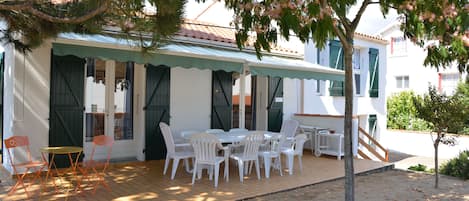 Un espace extérieur aménagé avec des sièges couverts, des chaises blanches et une table, un escalier menant à un bâtiment, et un arbre aux fleurs roses.