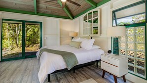 Une chambre à coucher comprenant un lit, une table de chevet, un ventilateur de plafond, une fenêtre donnant sur un espace verdoyant et une porte menant à l’extérieur.