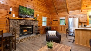 A log cabin interior with a stone fireplace, a television mounted above it, a dining area with a table and chairs, and a kitchen area with cabinets and appliances. - Stylish Cabin Near Pigeon Forge (Sevierville)