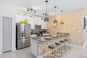 A modern kitchen with a central island, stainless steel appliances, pendant lighting, and a patterned wall.