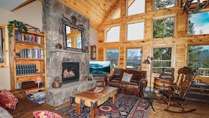 A cozy log cabin interior with a stone fireplace, wooden bookshelves, leather furniture, and large windows offering mountain views. - Grandview Chalet - Modern & Luxurious Mountain Retreat with Panoramic Views (Grand Lake)