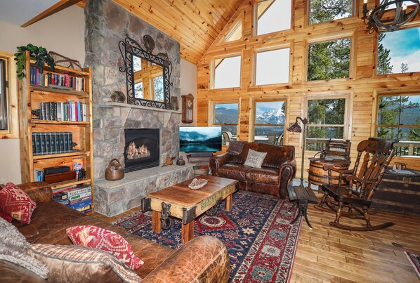 A cozy log cabin interior with a stone fireplace, wooden bookshelves, leather furniture, and large windows offering mountain views. - Grandview Chalet - Modern & Luxurious Mountain Retreat with Panoramic Views (Grand Lake)