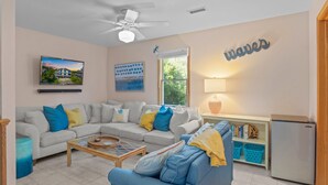 Ein Wohnzimmer mit grauem Sofa, blauem Sofa, Couchtisch, Deckenventilator, Fernseher, Bücherregal und Kühlschrank.