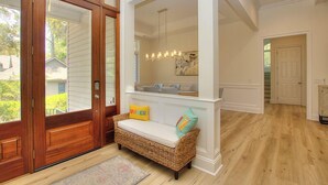 Un vestíbulo con puerta de madera, un banco de mimbre con cojines y una araña.