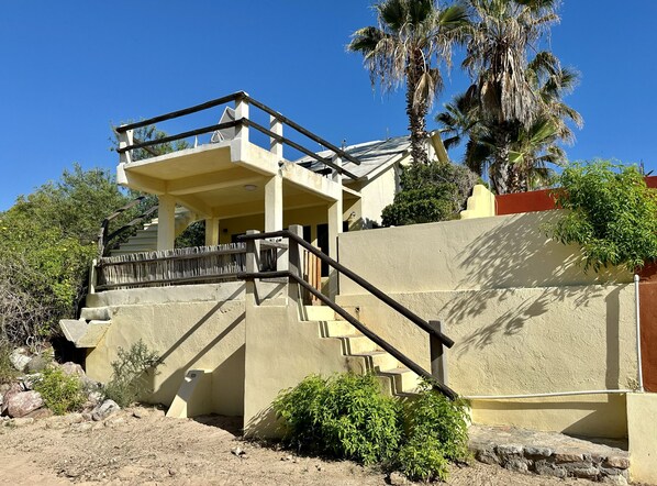 Un edificio de dos pisos con un balcón, una escalera y una palmera al fondo.