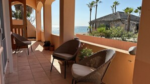 A balcony with wicker chairs and potted plants, overlooking a beach and a stone building.