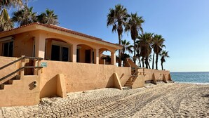 A beachfront property with a stucco exterior, a staircase leading to a covered area, and palm trees in the background.