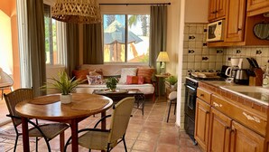 A cozy living area with a sofa, dining table, and chairs, a kitchen with wooden cabinets and tiled backsplash, and a large window with curtains.