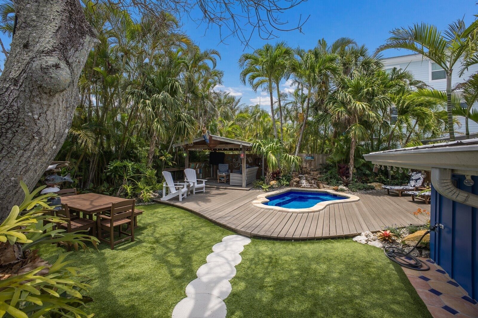 Un jardin avec une terrasse en bois, un jacuzzi et un espace salon abrité, le tout entouré de palmiers et d’une végétation luxuriante.
