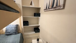 Ett litet, välordnat sovrum med en säng, ett litet bord med en växt, en hylla med förvaringslådor och en tavla på väggen.