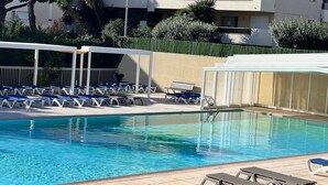 En swimmingpool med blå och vita solstolar, ett avskärmat sittområde och en byggnad i bakgrunden.