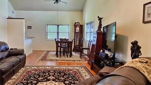 A living room with a leather couch, a dining table with chairs, a TV stand, and a large window with blinds. - The Haven of Peace in Madison County, FL (Lee)