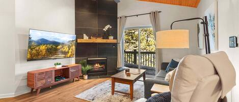 A living room with a fireplace, a television, a ceiling fan, and a view of trees through a window.