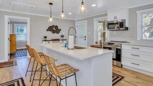A modern kitchen with white cabinetry, a central island with bar stools, stainless steel appliances, and pendant lighting. - Modern Waterfront Cottage on Wilmington by Lucky Savannah (Savannah)