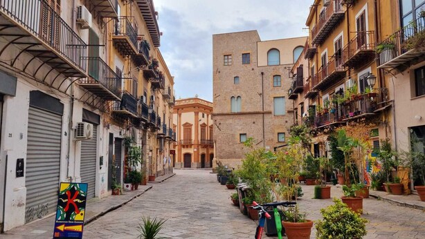 Una estrecha calle empedrada flanqueada por edificios de varios pisos, balcones con plantas en macetas y un edificio central con ventanales arqueados.