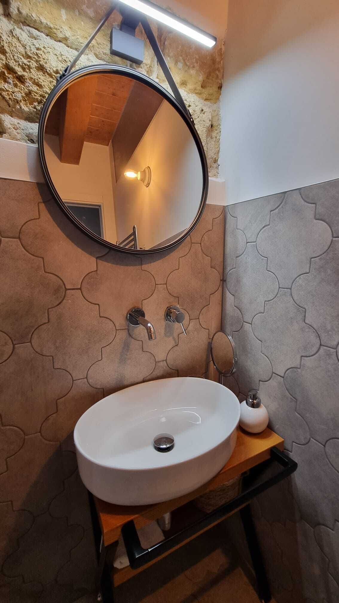 Een badkamer met een ronde spiegel, een witte wastafel en een handspiegel op een houten plank.