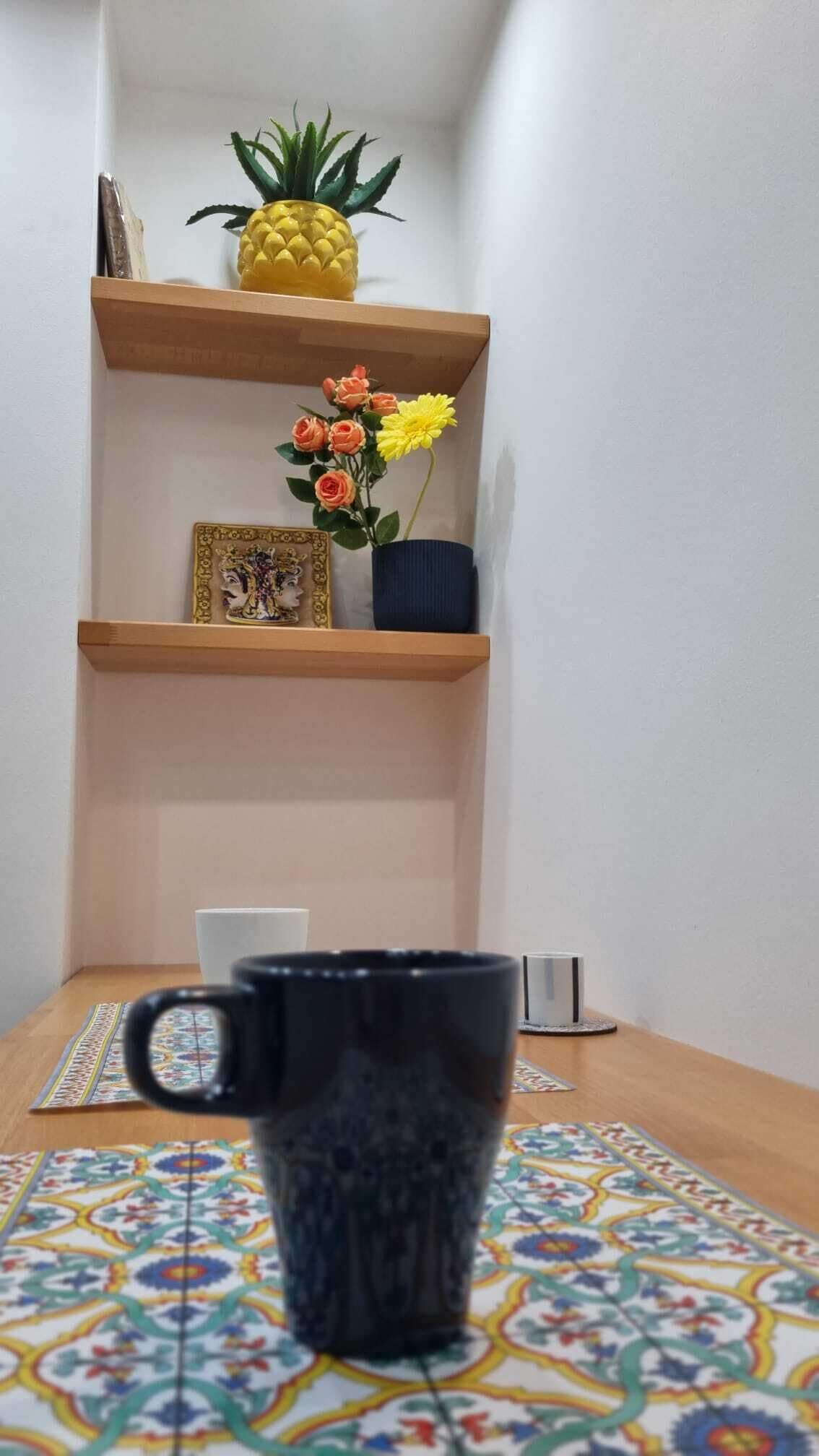 A corner of a room with a wooden shelf holding a pineapple, a vase with flowers, and a framed picture. A black mug is on a patterned tablecloth.