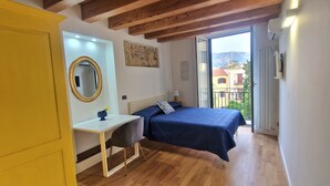 Un dormitorio con cama, escritorio, silla, espejo y balcón con vista a edificios y montañas.