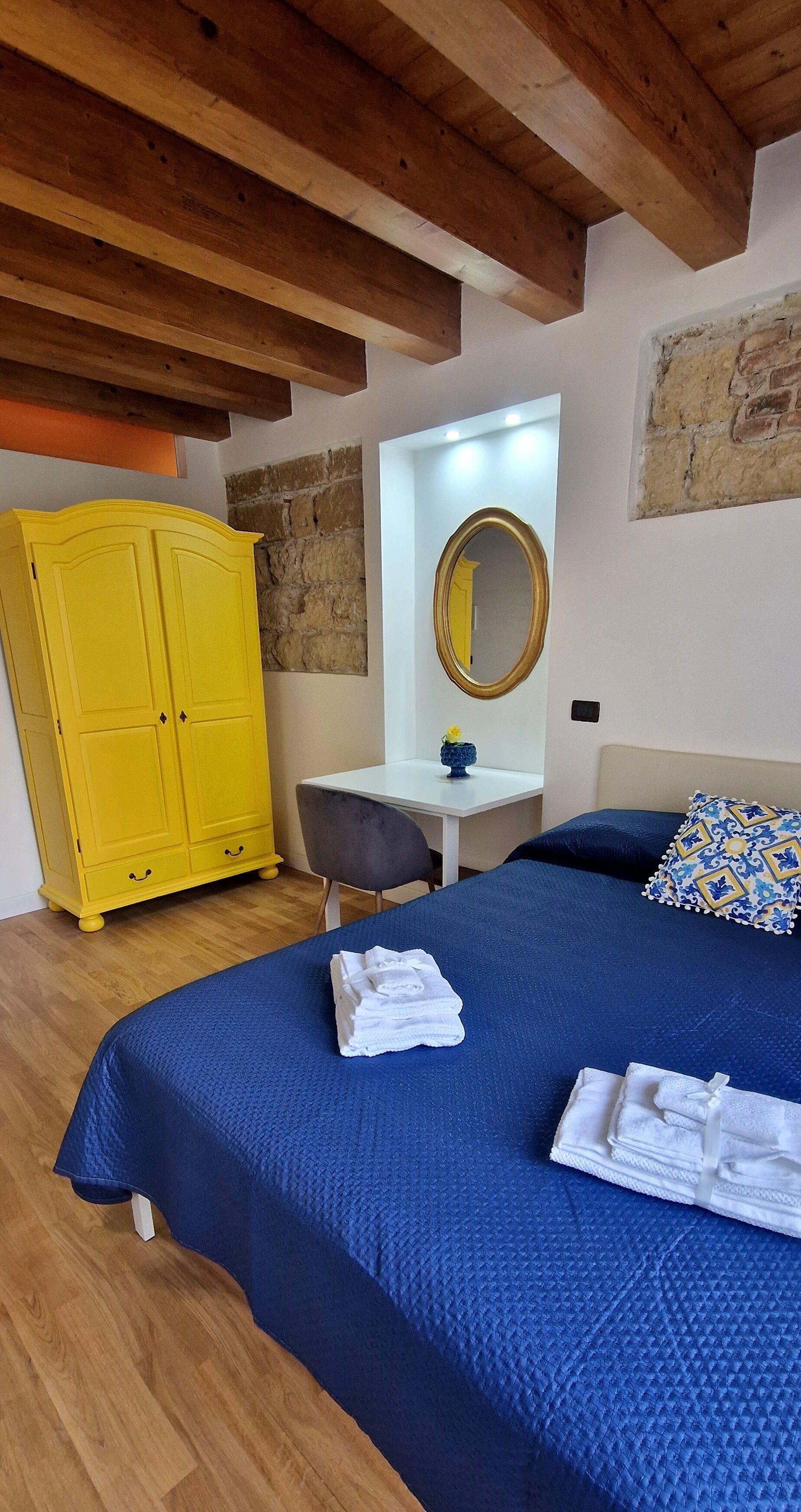 Une chambre à coucher avec un grand lit, une armoire jaune, un bureau avec un miroir et une couverture de lit bleue.