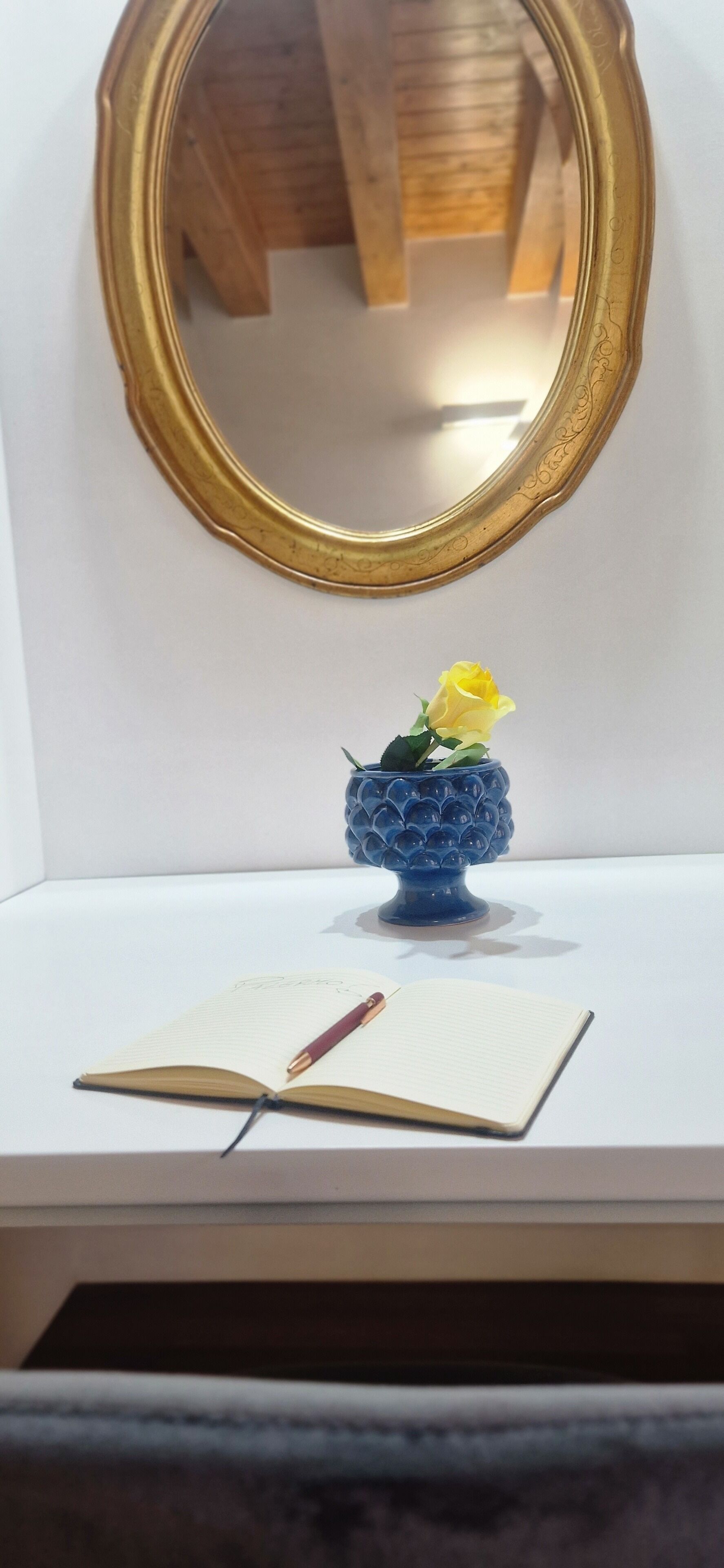 A white desk with a blue vase containing a yellow rose, an open notebook, and a pen.