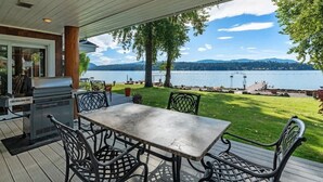 A covered patio with a table and chairs, overlooking a lake with mountains in the distance.