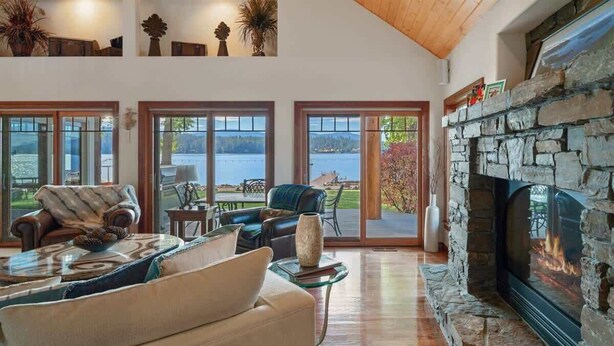 A spacious living room with a stone fireplace, large windows offering a view of a lake, and a cozy seating area with a sofa and armchairs.