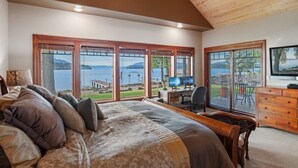A bedroom with a large bed, a desk with a computer, and a TV. There are large windows with a view of a lake and mountains.
