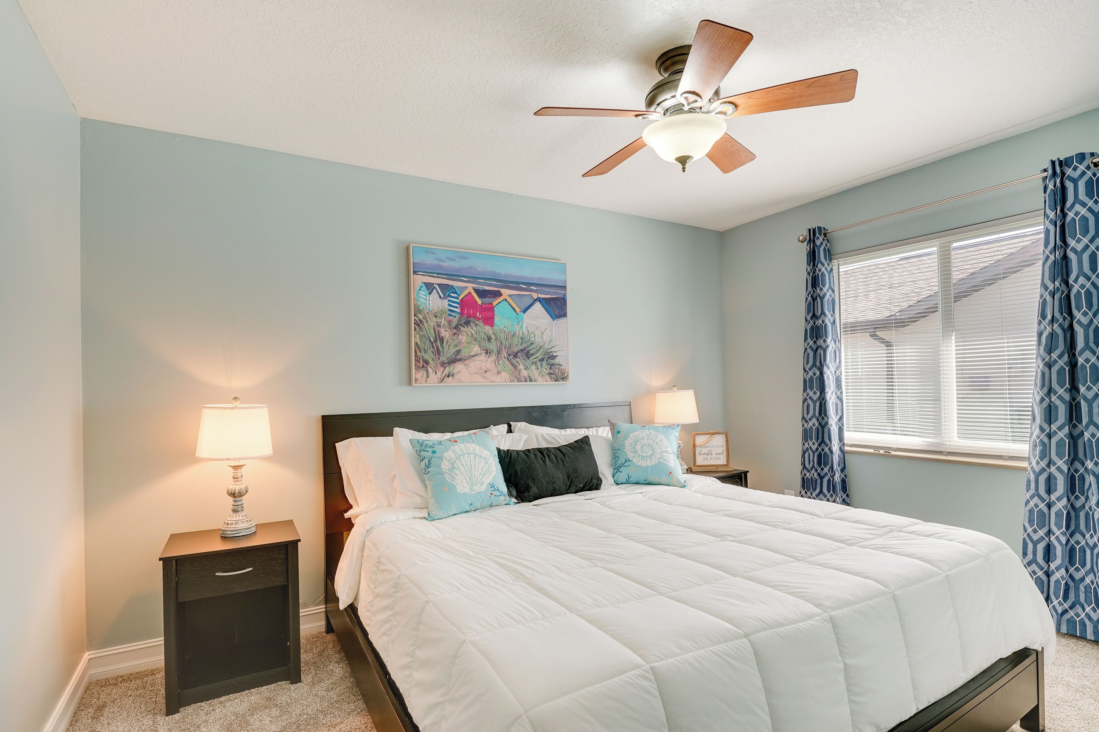 A bedroom with a bed, two bedside tables, a ceiling fan, a window with curtains, and a painting on the wall.